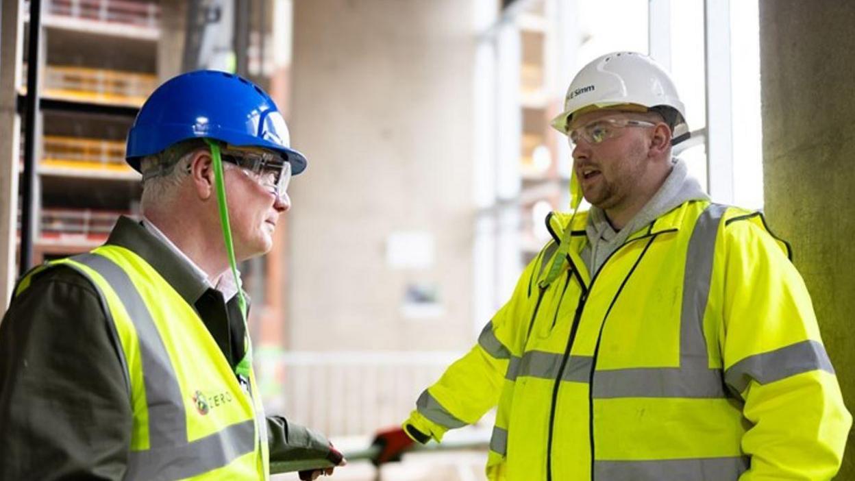 Two men in yellow reflective jackets and white hard hats in an empty concrete-walled building