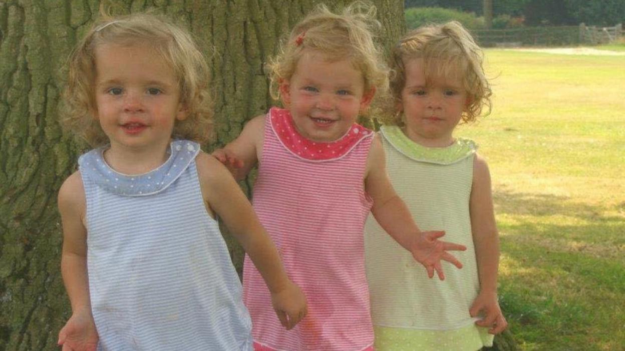  An image of the Orgill sisters as toddlers. Triplets Rose, Maudie and Grace Orgill are all wearing matching striped dresses in pastel blue, pink and yellow/green. They are standing at the base of a tree, smiling at the camera. They each have short blonde curly hair pinned up with a clip.