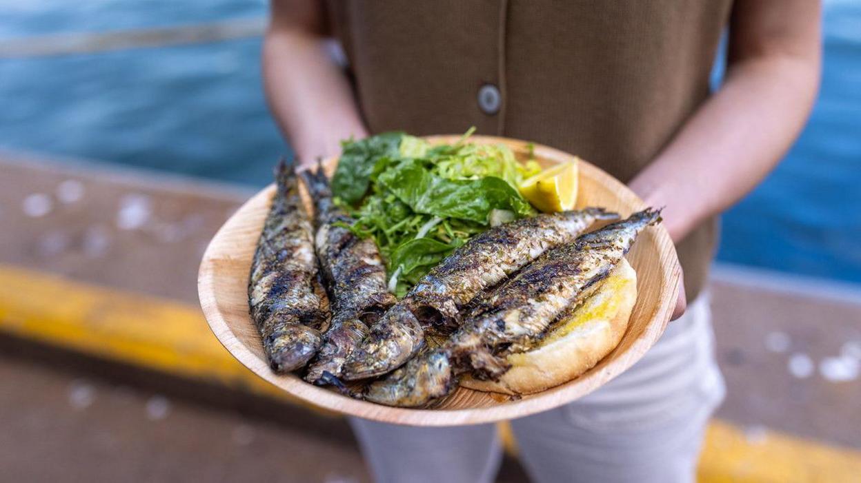 A person wearing a brown shirt and grey trousers stands on a harbourside with a plate of grilled sardines with a slice of bred and green salad.