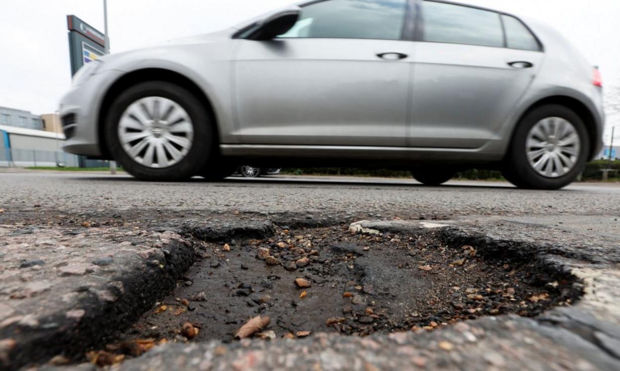 A pothole is in close-up at the bottom of the frame, as a silver four-door car with silver alloy wheels, passes it on a road.