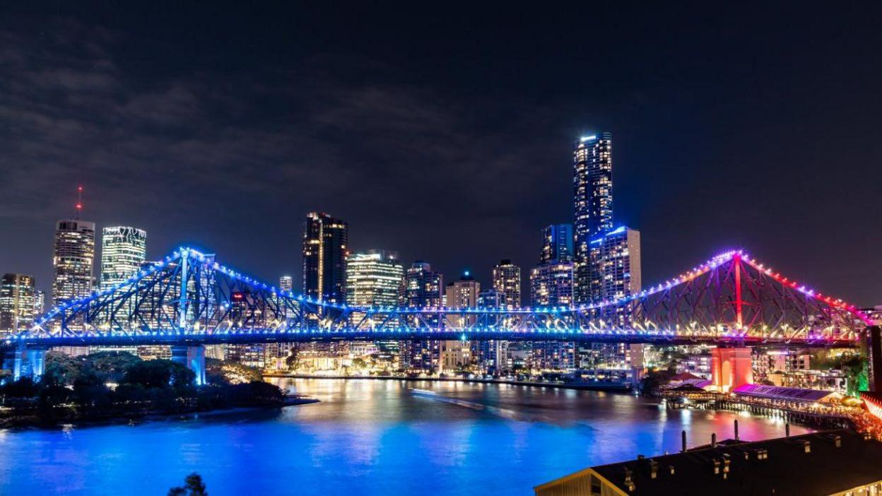 A road bridge going over a river lit up in the blue to red gradient of the climate stripes, underneath a night sky with high-rise buildings behind