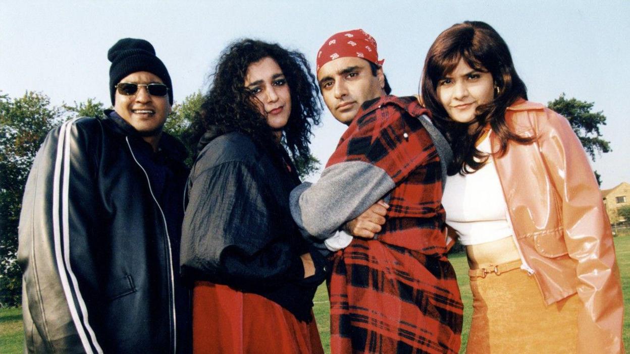 Four radical are lasting unneurotic connected a grassy tract with trees and a gathering down them. They are posed successful a consecutive line, each wearing different, brightly styled outfits.