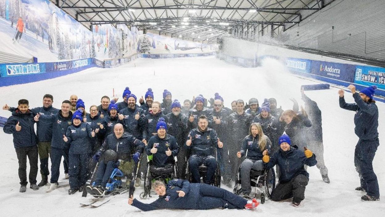 A group of people all smiling at the camera at the bottom of an artificial indooer ski slope. Some people are in wheelchairs. 
