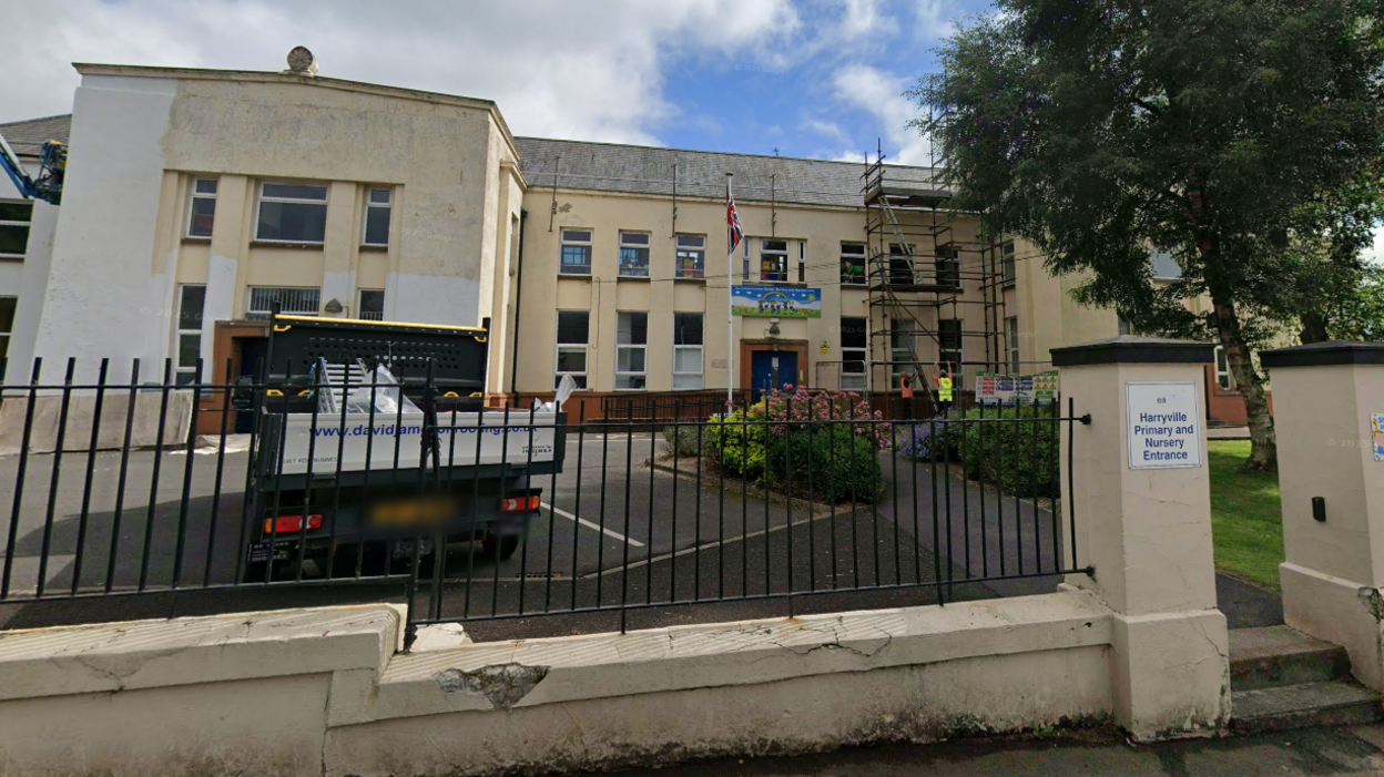 An exterior shot of a school. The school building, wall and gate pillars are yellow. The wall mainly consists of black bars. There is a tree and some bushes by a car park in front off the school. There is pickup truck parked outside. There is also a flagpole with a Union Jack and some scaffolding.