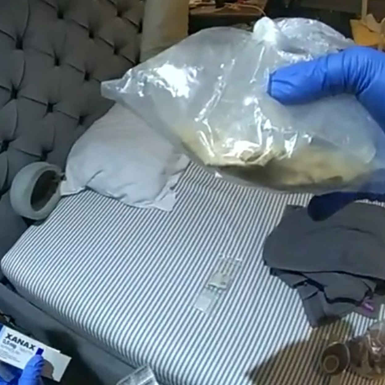 A police photograph inside Zou's flat. A bed can be seen and an officer's gloved hand is holding up a plastic bag containing a substance.