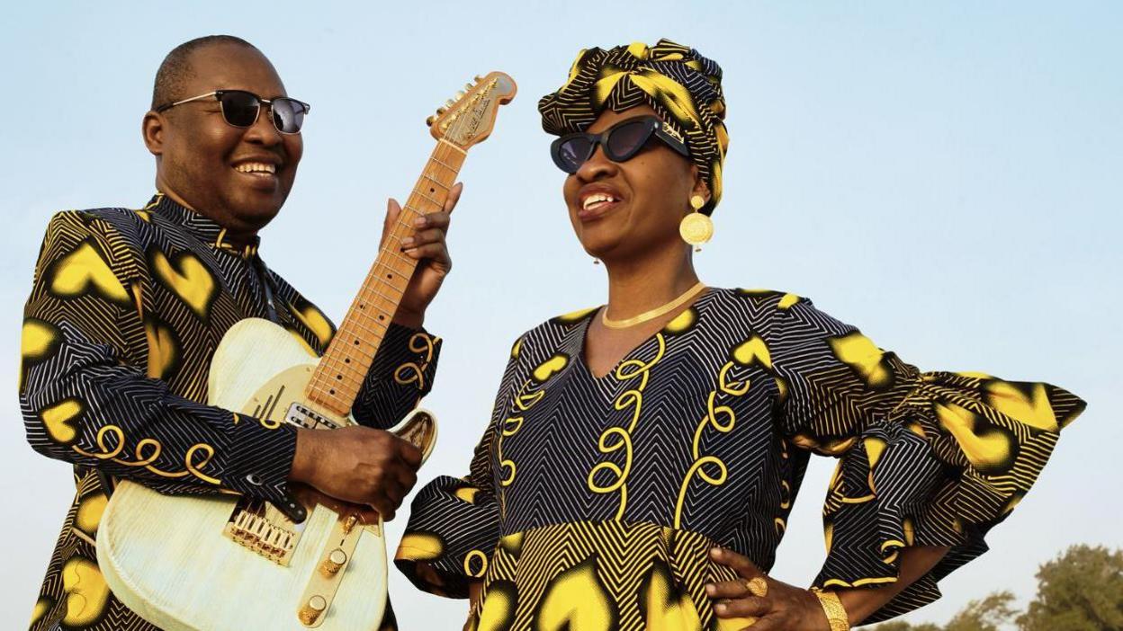 Music act Amadou and Mariam smiling. One man and one woman both in black and yellow outfits. 