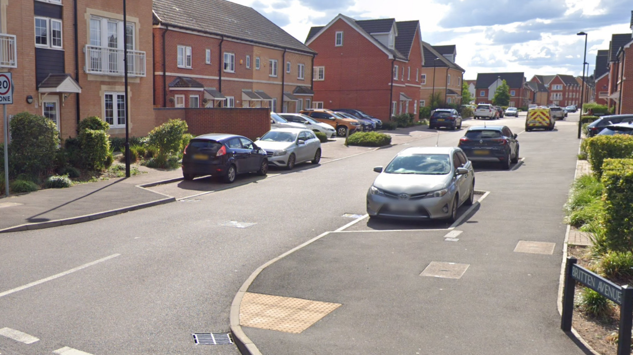 The street has what look like fairly new build homes. Parked cars are either side in bays just off of the road. There's a few streetlights. There's clouds in the sky but it's not overcast.