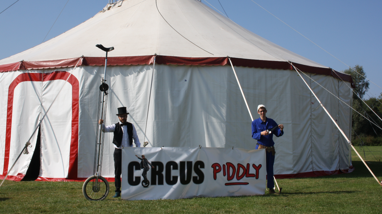 Sam Morley and Sam Goodburn standing in front of a white circus tent. Sam is holding a large unicycle, and they are standing in front of a sign saying 'Circus piddly'