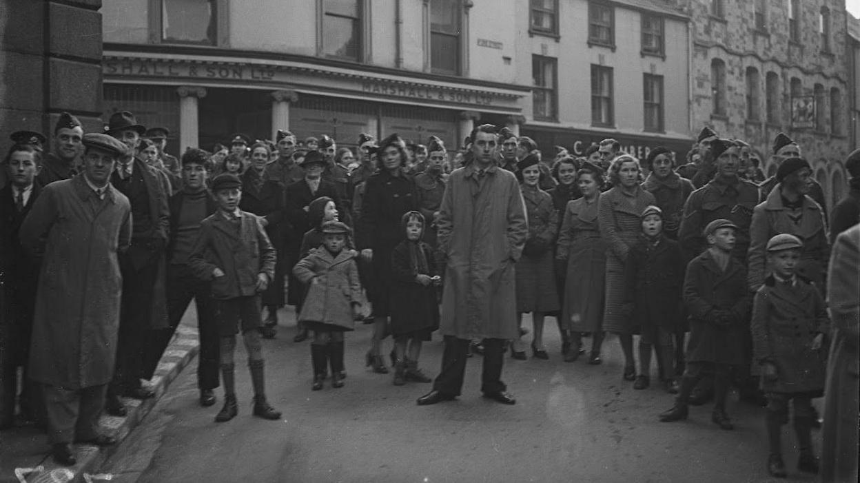 Black and white image of a crowd watching - some in military uniform - with building behind