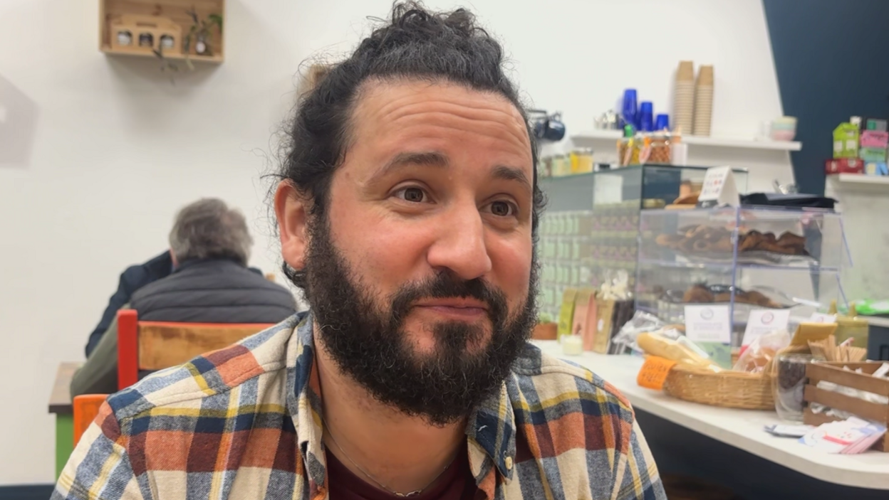 Giuseppe, speaking in a cafe. He has dark hair tied into a bun, and a dark beard. He has a checked shirt on and behind him is the cafe counter. `