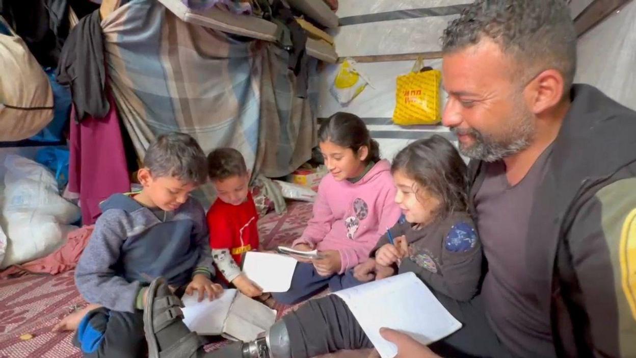 Man sits with four children on the floor of a shelter, with each of them holding some paper whilst one boy writes as they all look on