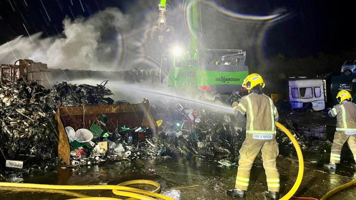 The scene is dark and two firefighters, one with a water hose, are extinguishing the metal that was on fire. Now smoke can be seen all around and a claw-like crane vehicle is in the distance 