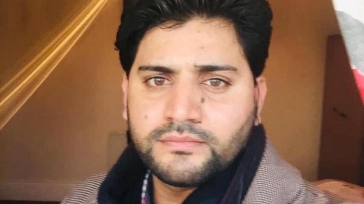 Shah has short thick black hair and a short stubbly beard. He is wearing a black scarf over a herringbone jacket and checked shirt