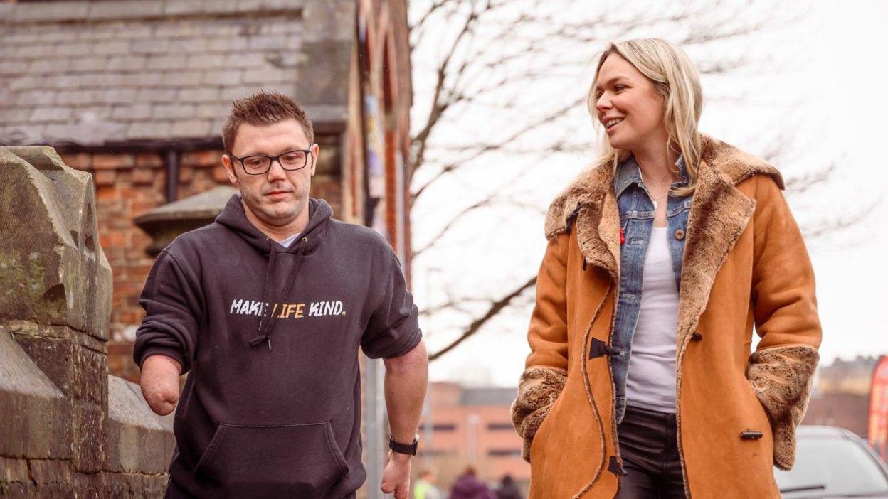 Lyndon and singer Hayley McKay chatting and smiling as they walk down the street. Hayley has medium-length blonde hair. She is wearing a denim shirt, white top and black trousers. Her hands are in the pockets of her brown coat. Lyndon is wearing a black hoodie reading Make Life Kind and black glasses.