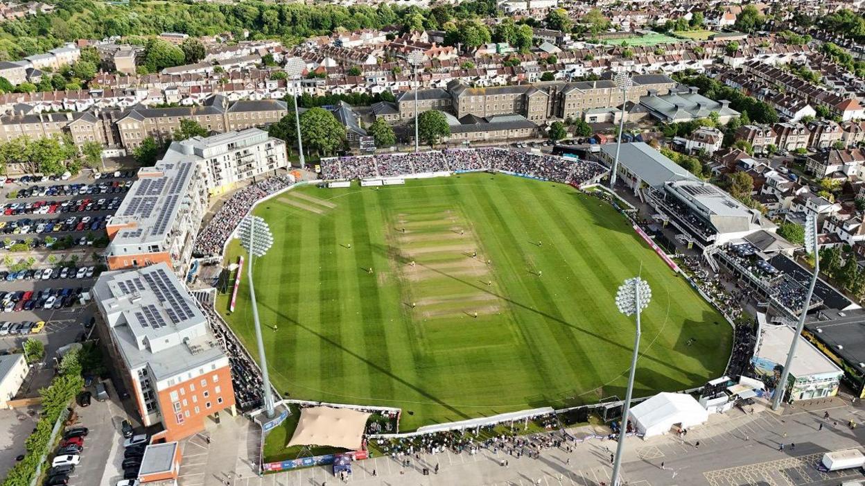 An aerial shot of the County Ground in Bristol during a T20 cricket match. Temporary stands full of spectators are visible, as are the high-rise flats that overlook the stadium. It is a sunny evening with the floodlights casting shadows across the pitch. The image is taken from a drone