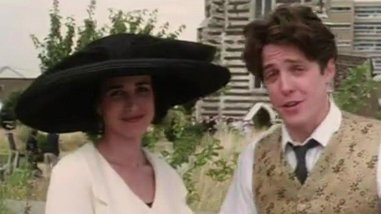 A woman (Andie MacDowell) wearing a large black hat and a white suit jacket standing next to a man (Hugh Grant) wearing a white shirt, black tie and golden embellished suit vest. They are standing outside grasslands and a tall building to the right.