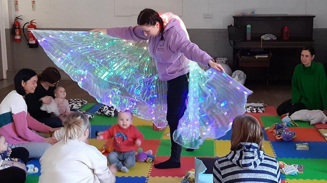 A young woman with brown hair that has been plaited is wearing large multi-coloured fabric wings with lights attached, as well as a purple hoodie and black leggings and socks. She is waving the wings close to a baby who is sat on the floor during a baby sensory session. The baby is wearing a red jumper with blue jeans. Other babies and mothers sit in a circle around the baby and woman looking on. They are all sat on a colourful mat inside a hall.