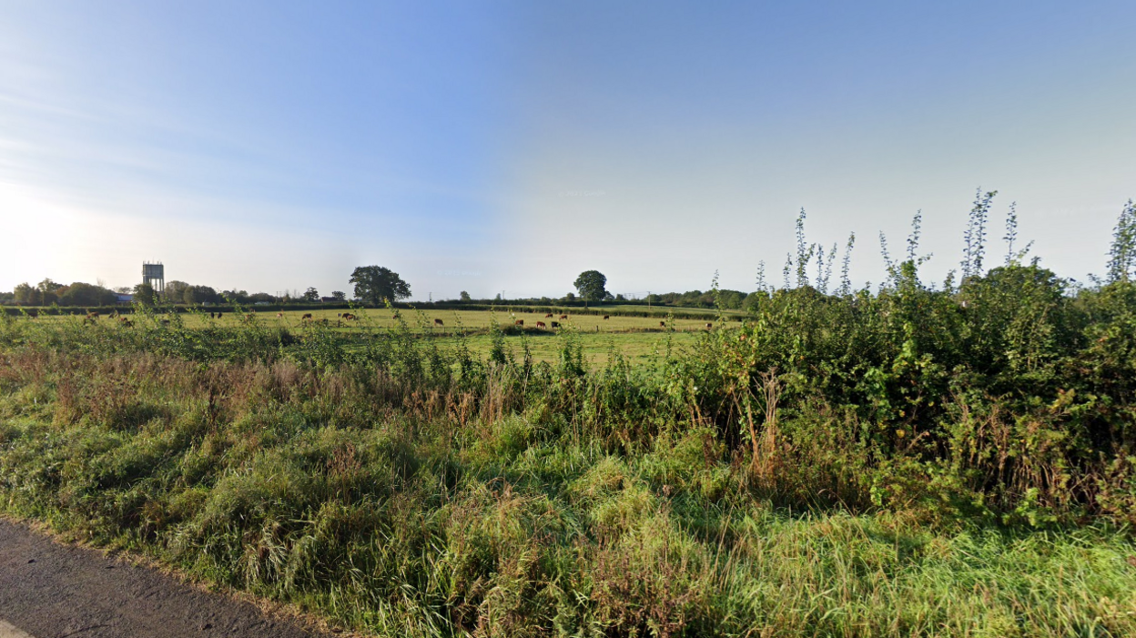 road in foreground with hedges and rolling fields with trees further back. Water tower off to the left.