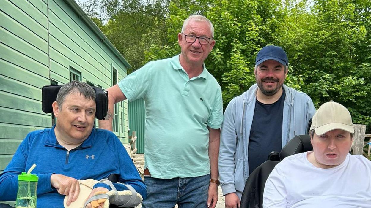 Alec is wearing a turquoise polo shirt, stood next to Dave who is wearing a grey hoodie undone with a black t-shirt and blue hat. They are stood behind Sion and Shane who are wheelchair users. The group are pictured outside with trees in the background and stood next to a sage green wooden building.