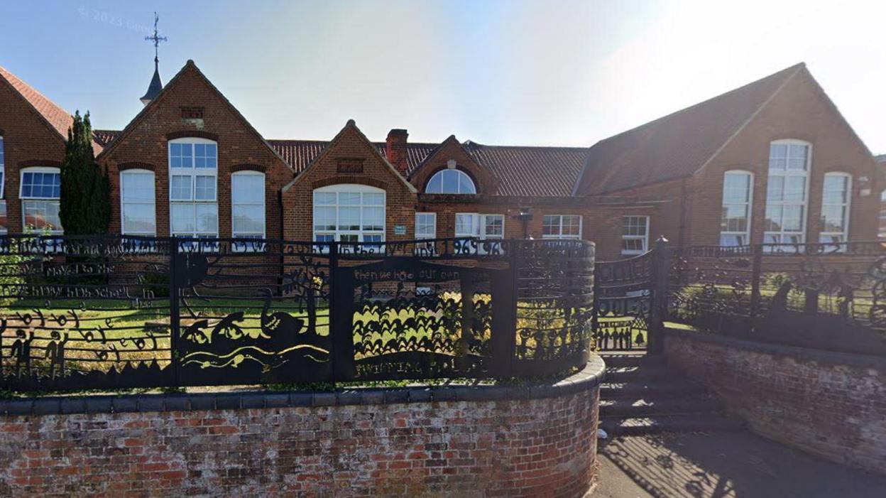 Black fences surround Fakenham Juniors, with grass in the foreground of the red brick building.