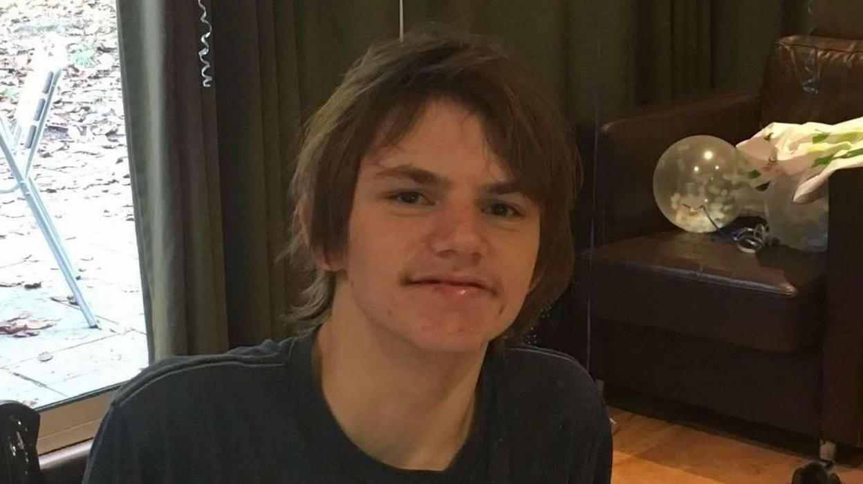 A man/teen is in a room with a leather chair behind. He has dark hair and is wearing a dark top. He is smiling. There's a big glass door behind looking into a garden.