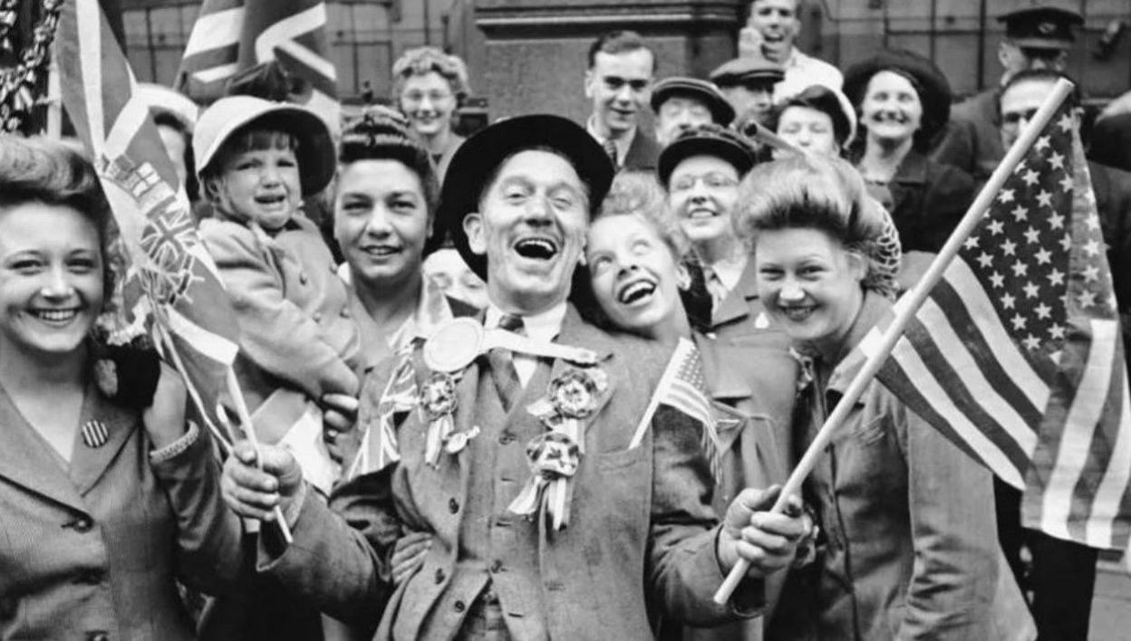 A black and white image of a group of adults and children of varying ages waving flags and smiling at a party to celebrate VE Day in 1945 
