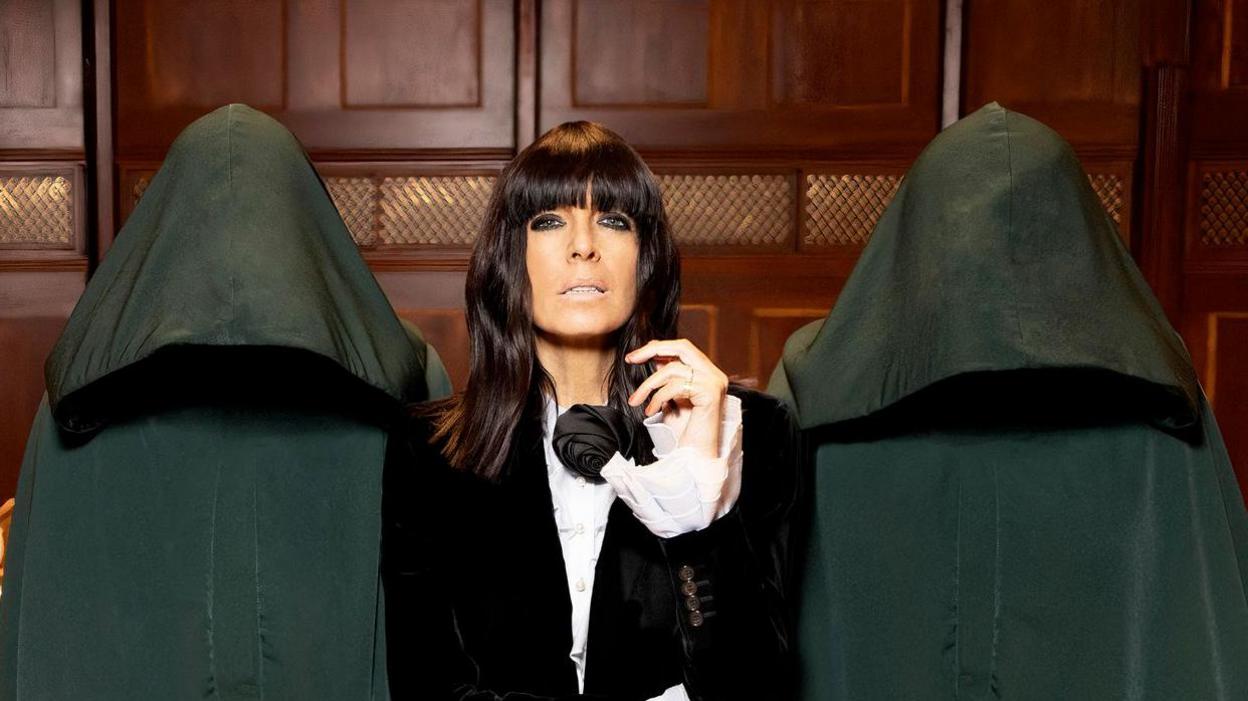 Claudia Winkleman wears a dark suit and white shirt. She has brown hair that goes just past her shoulders and wears heavy eyeliners. She is flanked by two anonymous people in hooded cloaks. They are in a wood-panelled room