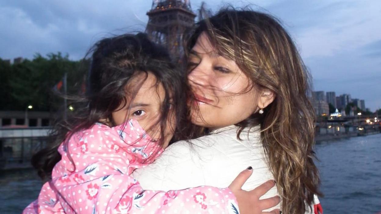 Mariam Kalim is wearing a pink poka dot floral coat holding her mother Zobia Kalim. They are both smiling in front of the Eiffel tower. 