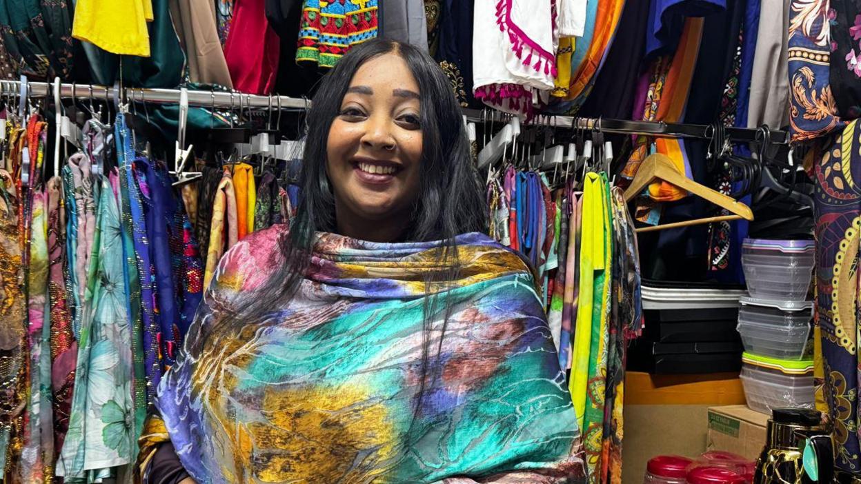 Athar Alsadek wearing colourful clothing. She is in a market and there are clothes on a rail behind her. She has long black hair and is wearing a smile. 