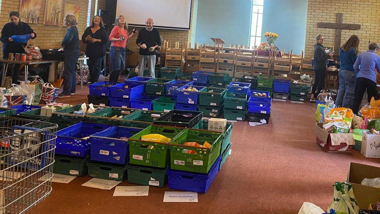 Lost of plastic food containers being stacked in a church. The boxes are green and blue and you can see food items such as cereal sticking out of the top.  You can see volunteers in the back of the image. 