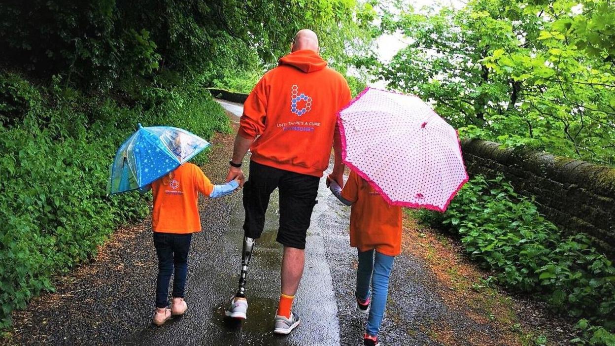 Pete Lloyd walking down a country path holding hands with two of his children. They all have their backs to the camera - the children are holding umbrellas. Pete is wearing a red Bone Cancer Research Trust hoodie and his prosthetic leg is visible. 