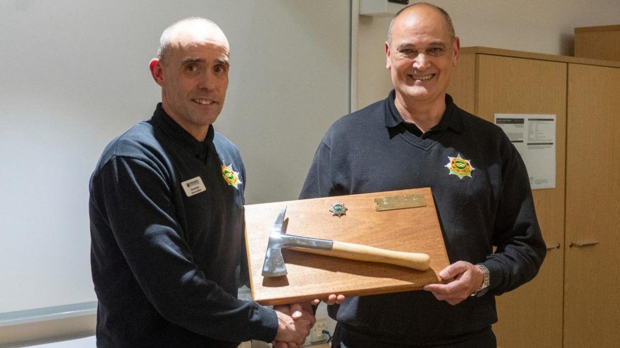 Two men wearing black tops with a Staffordshire Fire and Rescue Service logo shake hands as one passes the other a wooden board with a fire axe attached to it.