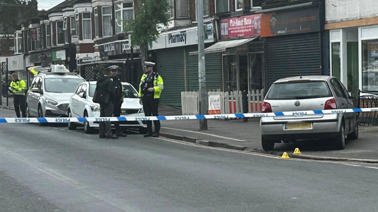 A silver VW Polo on the pavement in front of a row of shops. There is a police cordon in place, with tape across the road.