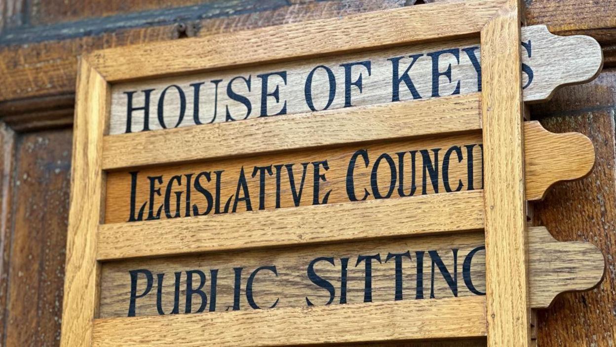 A wooden sign on the outside of the door to the Tynwald buildings. It has slats with the House of Keys, Legislative Council and Public sitting slotted into it.