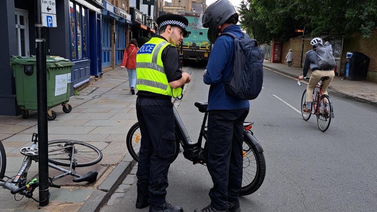 A police officer faces away from the camera, talking to a man wearing a black motorbike helmet and standing next to a black e-bike. They are on a city street with industrial bins and a bike tied up to a lamppost, with another cyclist riding past on a normal bike. The man is wearing blue clothes and has a blue rucksack.