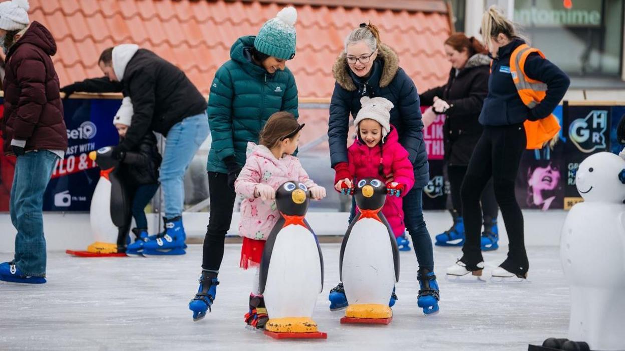 Ice Cube skating rink, with children and adults skating - the children hold on to penguin statues while they skate 