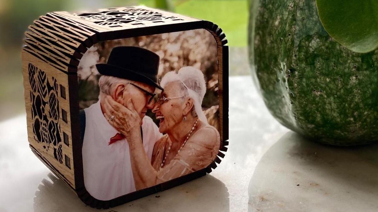 A wooden cube with round edges sits on a white side table. On the face that points forward is a picture of an old woman and an old man - both white - smiling and laughing. 