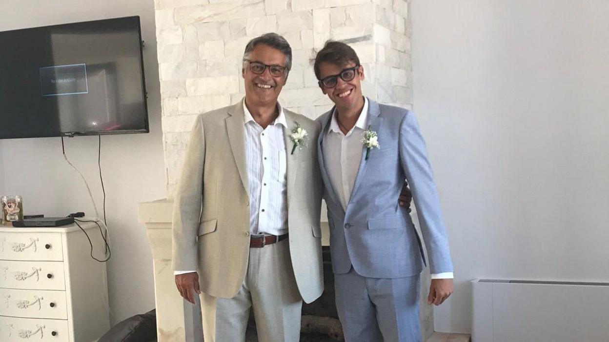 Nicolas Gerasimidis and his son Adam, both wearing light-coloured suits with flowers on lapels, smiling at the camera