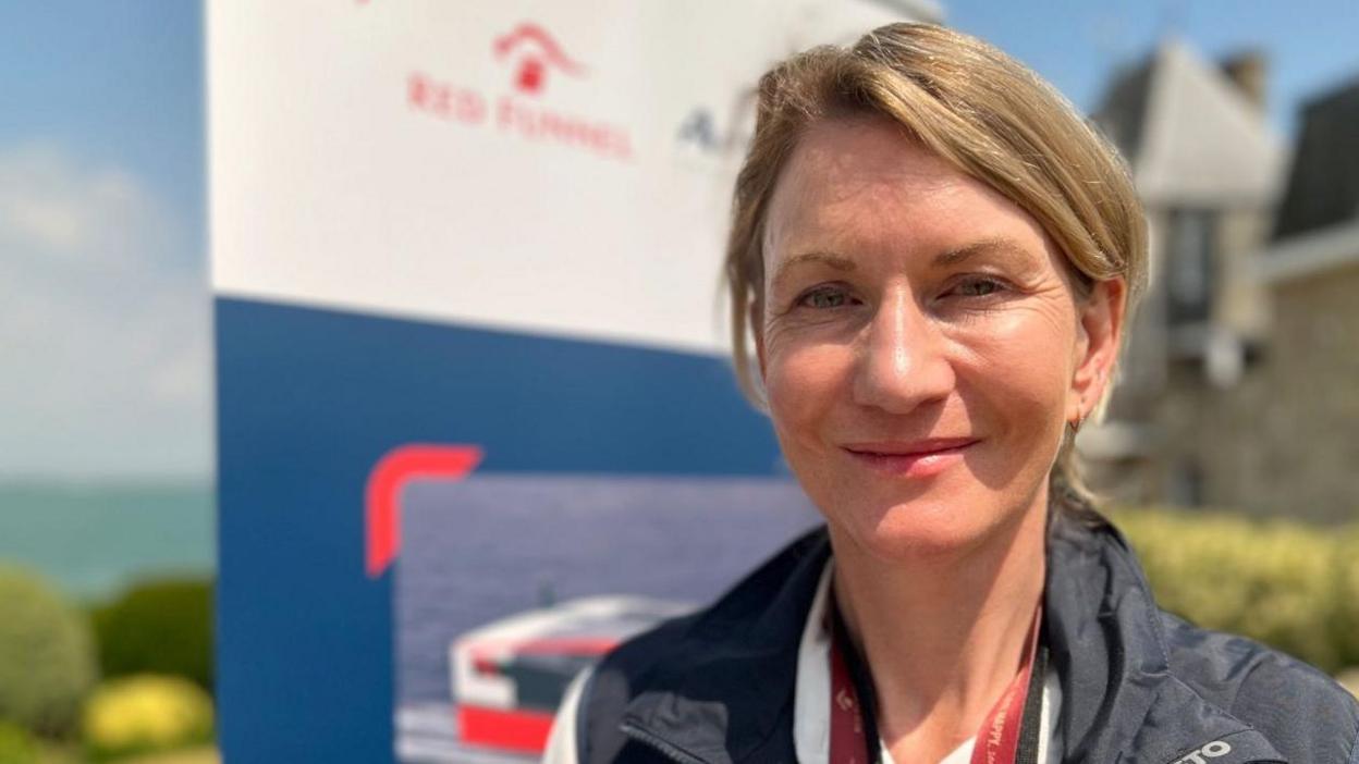 Fran Collins is slim with dark blonde hair. She is wearing a red lanyard around her neck and a dark navy jacket over a shirt. The background is out of focus, but shows a banner reading 'Red Funnel', with bushes and the sea further in the background.