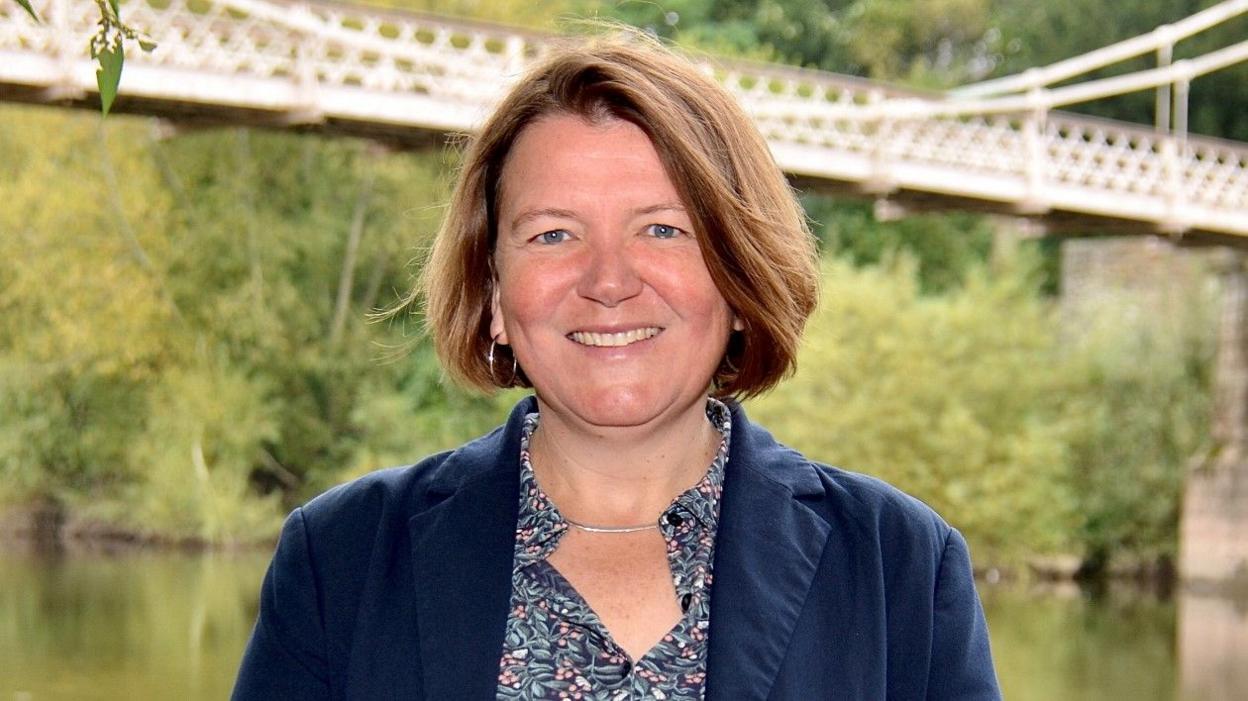 MP Dr Ellie Chowns smiling in front of the Victoria Bridge over the River Wye 