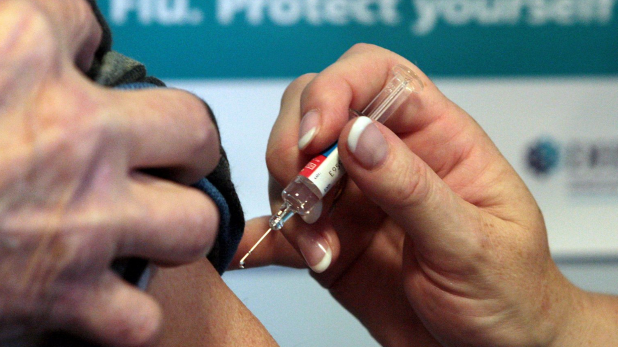 The image shows a close-up of a person receiving an injection in the upper arm. The syringe contains a clear liquid, and the person administering the injection is holding the needle and vial with both hands. The background includes a blurred sign with the words Flu. Protect yourself.