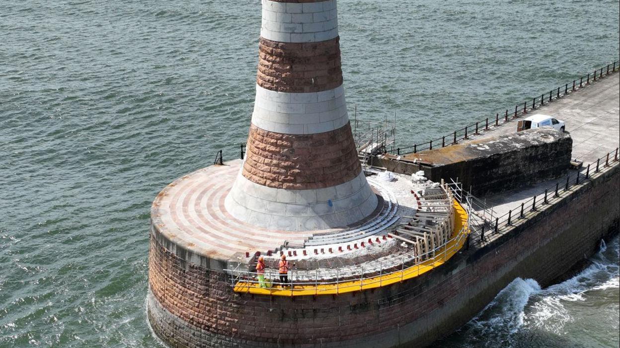 A white and brown lighthouse sits on top of a circular pier. Two people wearing hi-vis orange jackets and helmets can be seen on scaffolding on the side of the circular pier.