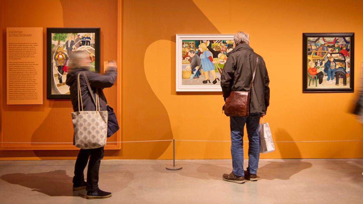 The photo shows people looking around the Beryl Cook: Pride and Joy exhibition, which ran between January and May. There is a bright orange wall on which three pieces of Beryl Cook art are hung. One woman is taking photos.