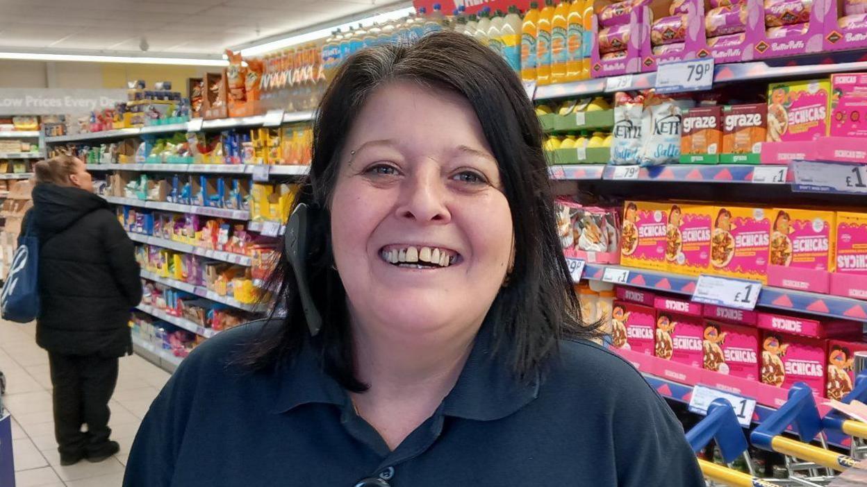 A woman with black hair and wearing a headset is smiling at the camera in a supermarket. 