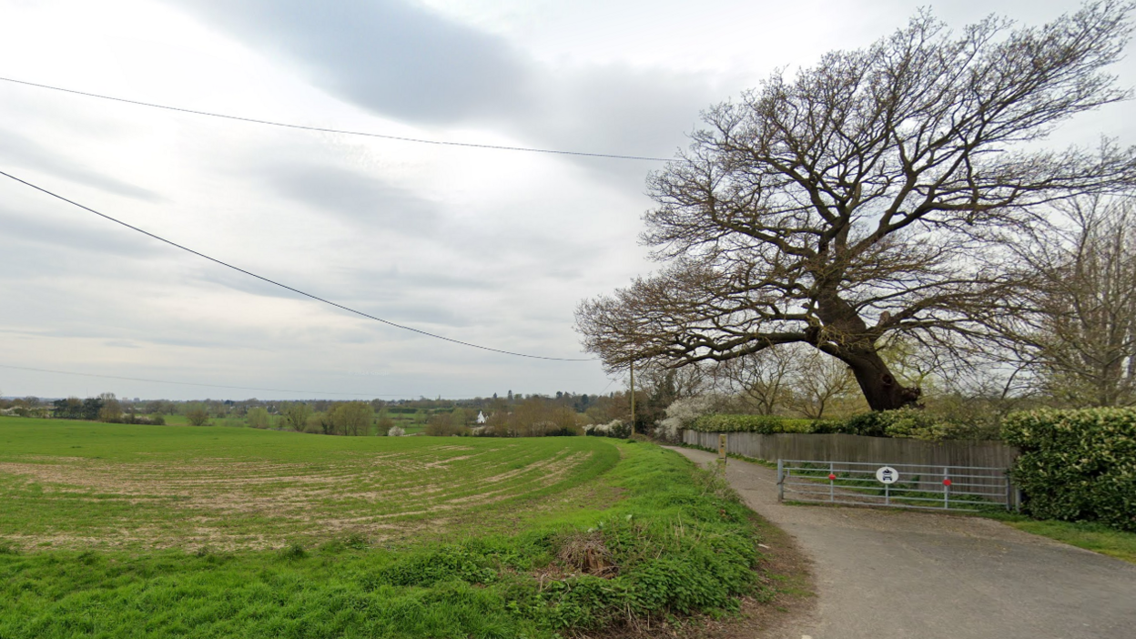 A view across the field at Mill Lane in Chelmsford. There is a metal fence in front of a path to the right, with the large field on the left.