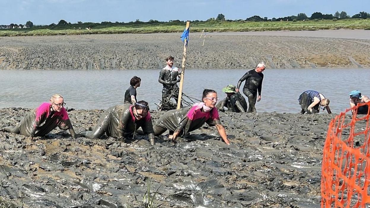 Three women crawling through the mud with five people and the river behind them, Maldon