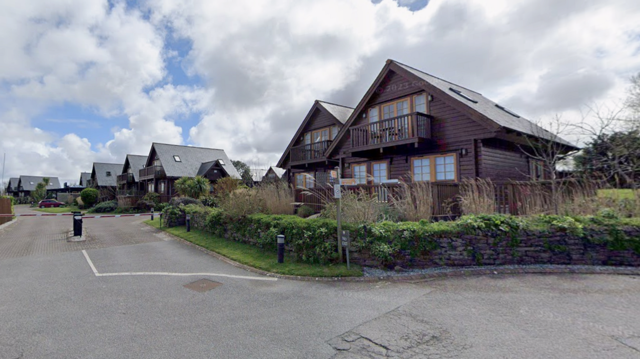 Several wood clad holiday lodges have pitched roofs, seats on the veranda and a balcony can be seen on the first floor. They are surrounded by shrubs.