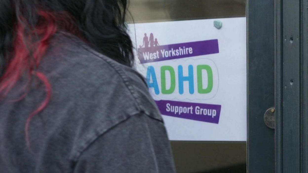 A person with red and black hair and a black jacket, with their back to the camera, faces a white sign attached to a door. The sign says, in colourful writing, West Yorkshire ADHD Support Group.
