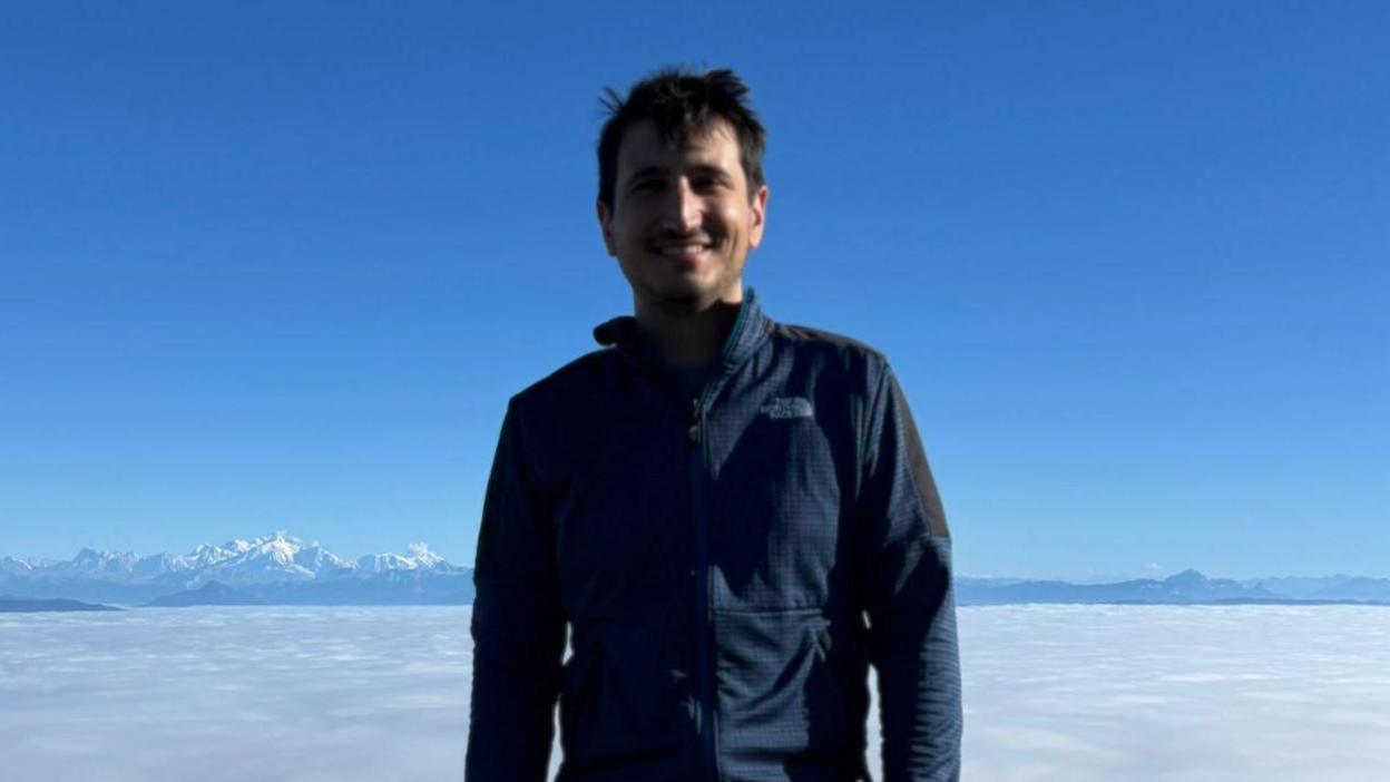 Federico is smiling with a blue sky in the background. He appears to be standing somewhere very high up, with mountains in the background, and clouds below him. He wears a blue outdoors jacket.