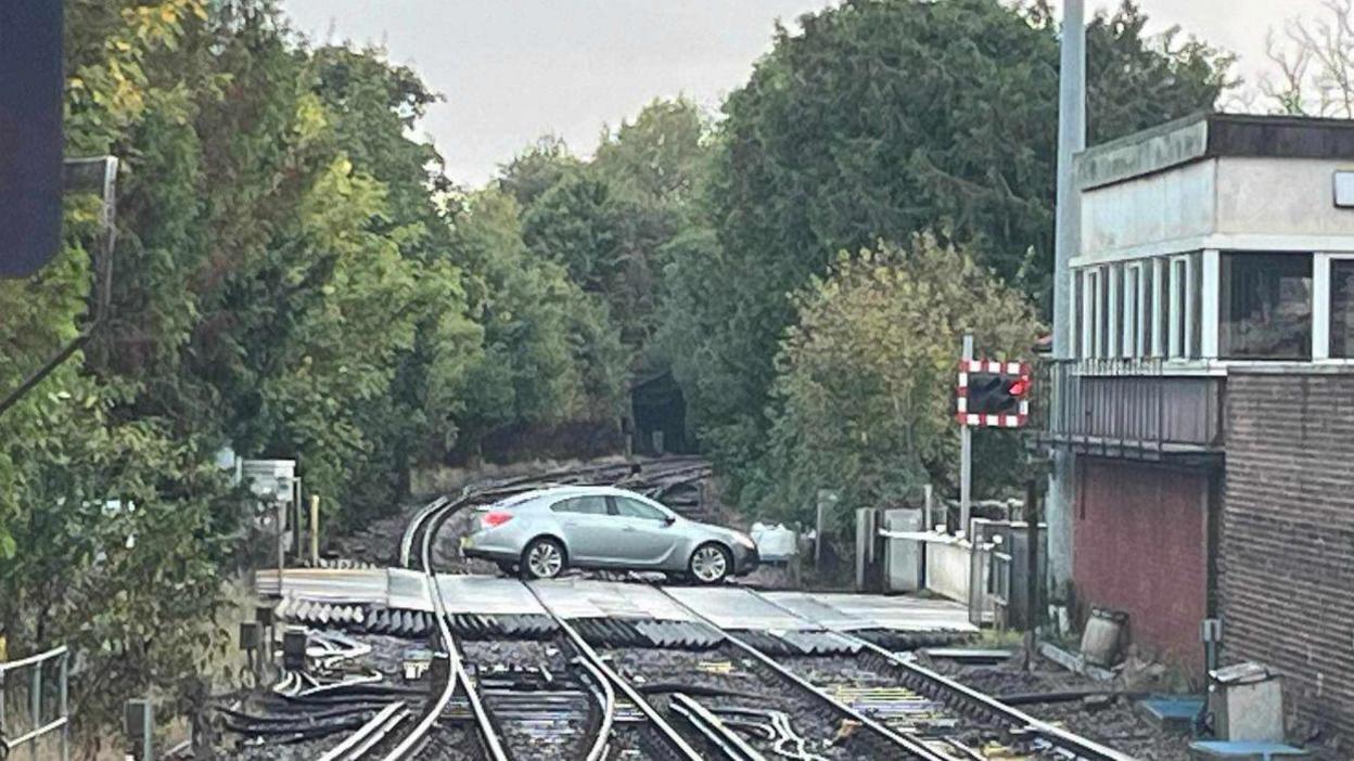 A photo of the car on the level crossing taken from further away. A railway building can be seen on the right hand side.